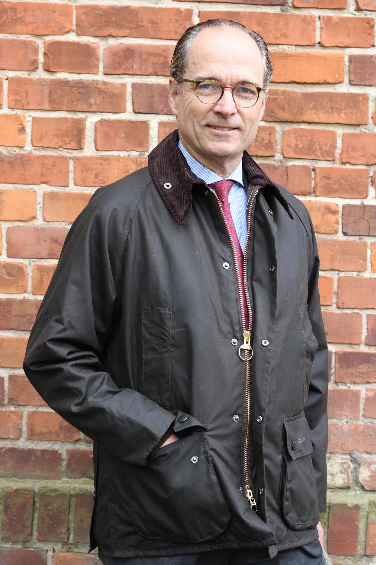 Bernhard Roetzel in der Barbour Classic Beaufort vor einer Backsteinwand, die Hand in der Tasche