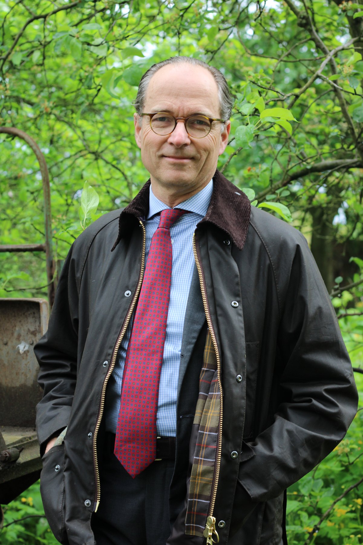 Bernhard Roetzel in der Barbour Classic Beaufort mit offenem Reißverschluss, im Futter das Tartanmuster, vor grünem Laub