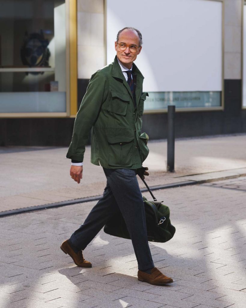 Bernhard Roetzel in a dark green field jacket and flannel trousers, carrying a green weekender bag.
