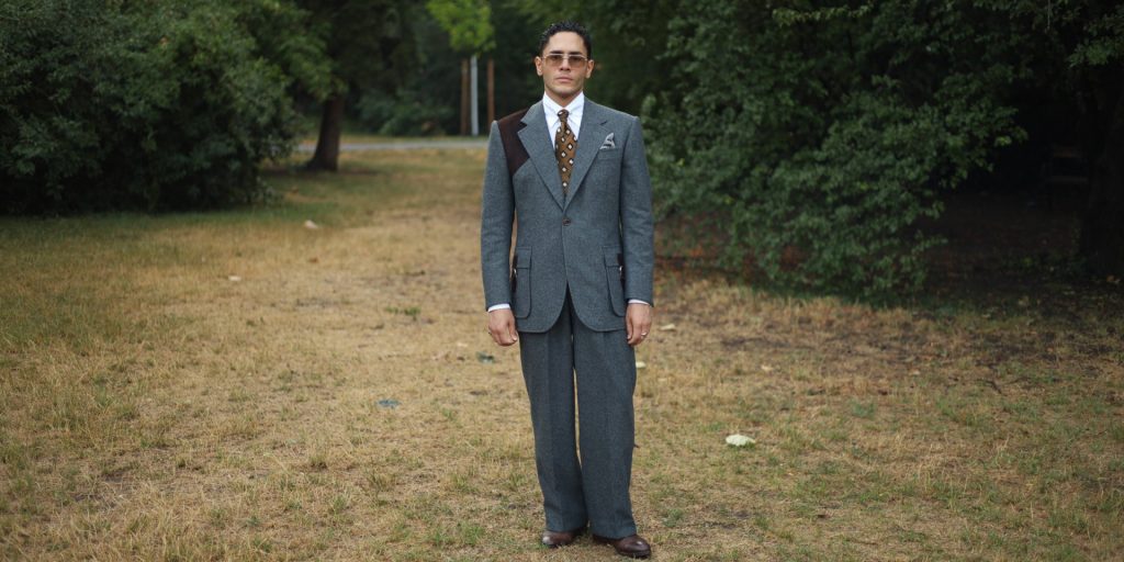Yasin Tarabia in einem graublauen Anzug mit weiter Hose, ganze Figur in einem Wiener Park