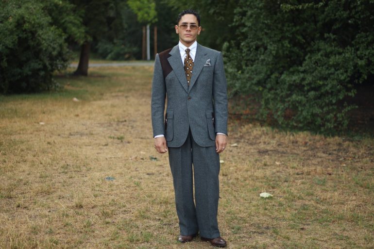 Yasin Tarabia in einem graublauen Anzug mit weiter Hose, ganze Figur in einem Wiener Park