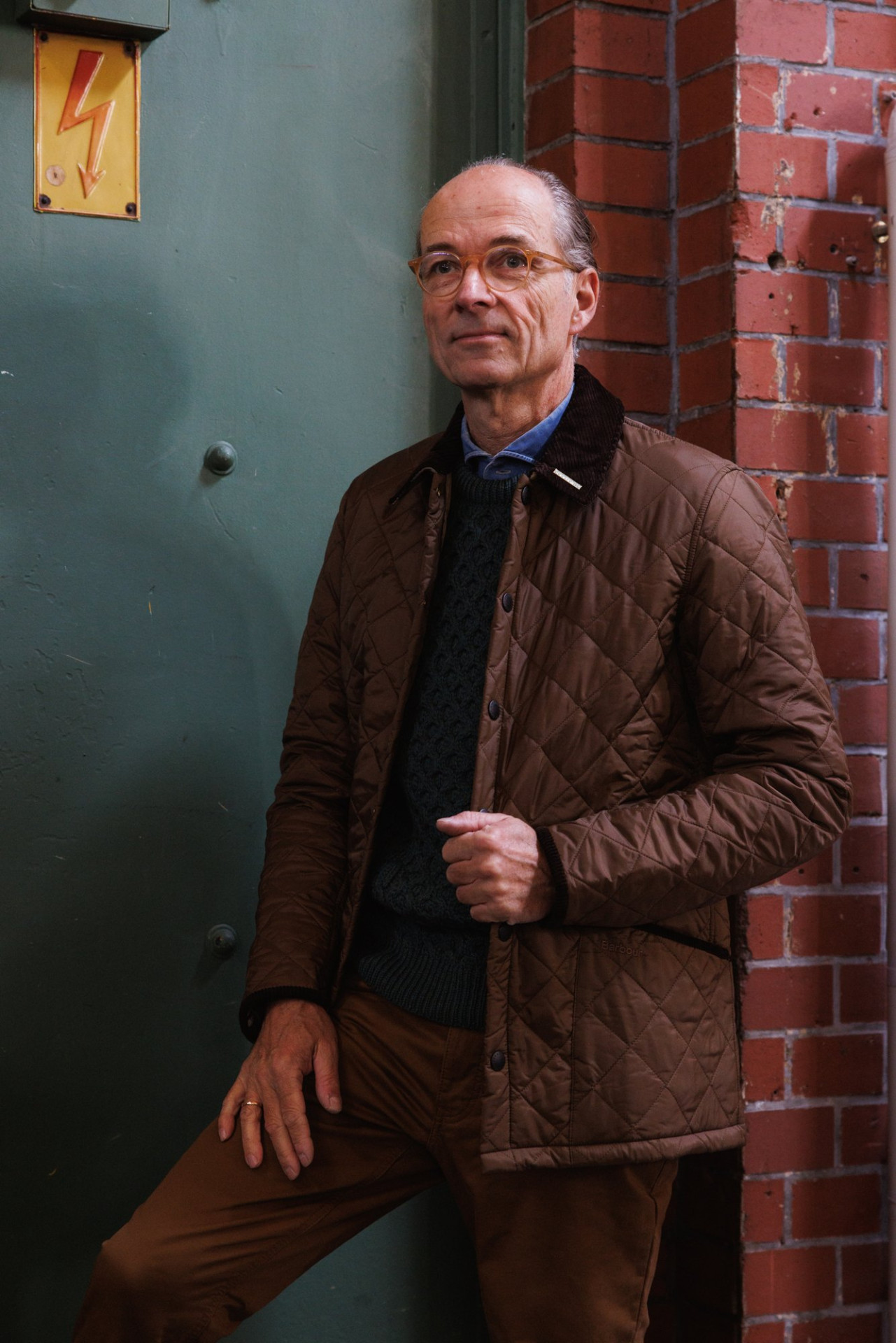 Bernhard Roetzel in der Steppjacke und dem grünen Aran-Pullover vor einer grünen Wand neben roter Backsteinmauer