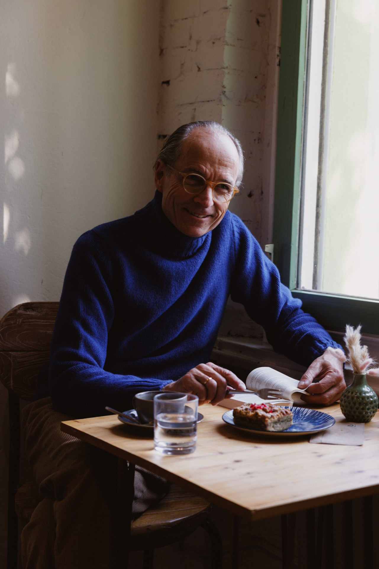 Bernhard Roetzel lächelt im blauen Turtleneck am Fenstertisch im Café, vor ihm ein Stück Kuchen