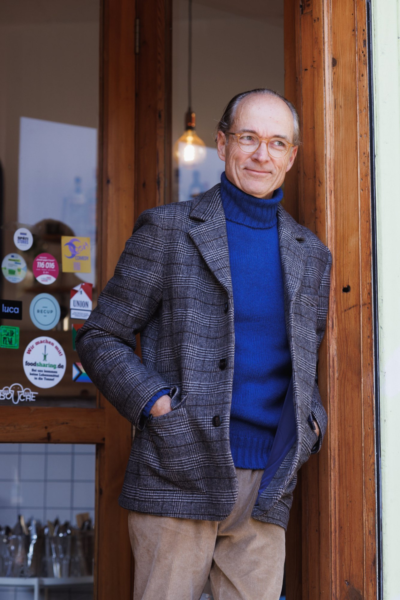 Bernhard Roetzel im Sakko mit Prince-of-Wales-Check, blauem Turtleneck und beiger Kordhose in einer Holztür mit Aufklebern
