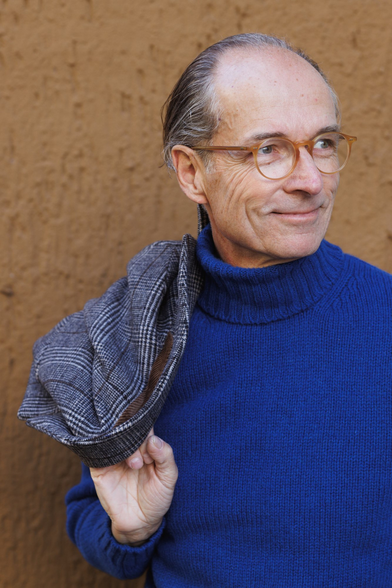 Bernhard Roetzel lächelt in die Kamera, in der Hand eine Mütze im Prince-of-Wales-Check, dazu ein blauer Turtleneck