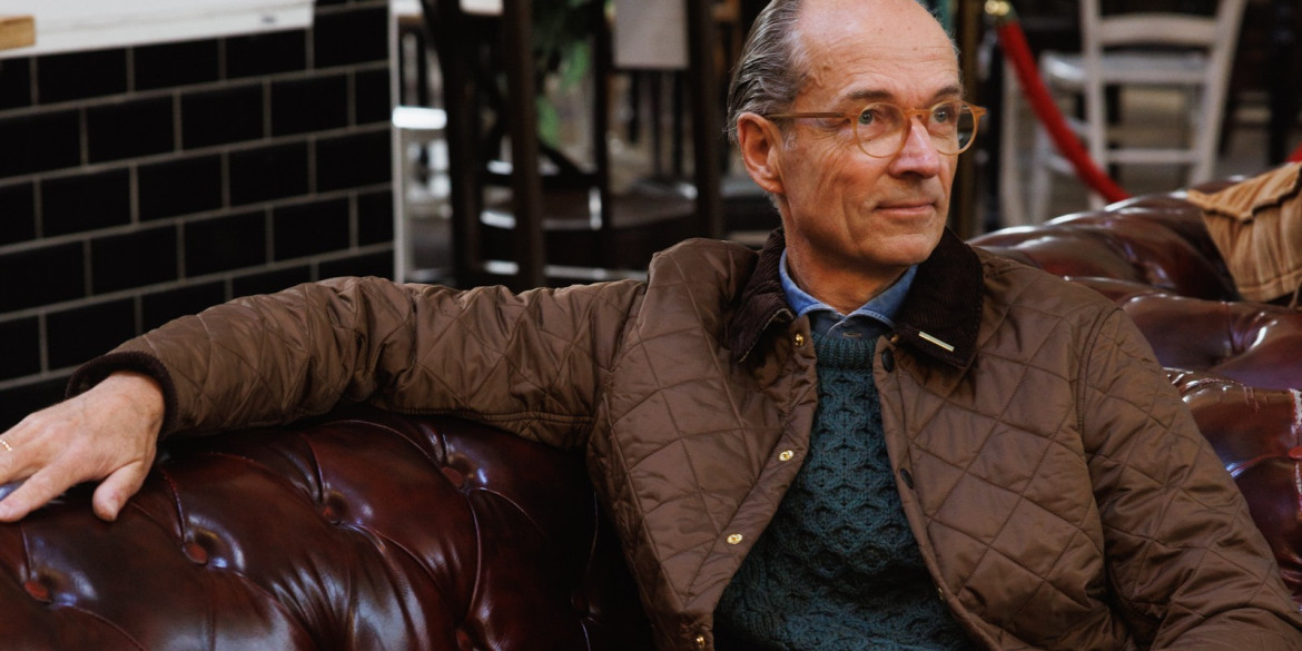 Bernhard Roetzel in gesteppter Jacke und grünem Aran-Pullover auf einem Chesterfield-Sofa im Café