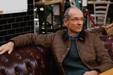 Bernhard Roetzel in gesteppter Jacke und grünem Aran-Pullover auf einem Chesterfield-Sofa im Café