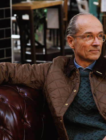 Bernhard Roetzel in gesteppter Jacke und grünem Aran-Pullover auf einem Chesterfield-Sofa im Café
