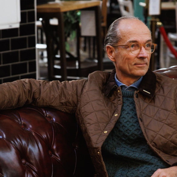 Bernhard Roetzel in gesteppter Jacke und grünem Aran-Pullover auf einem Chesterfield-Sofa im Café