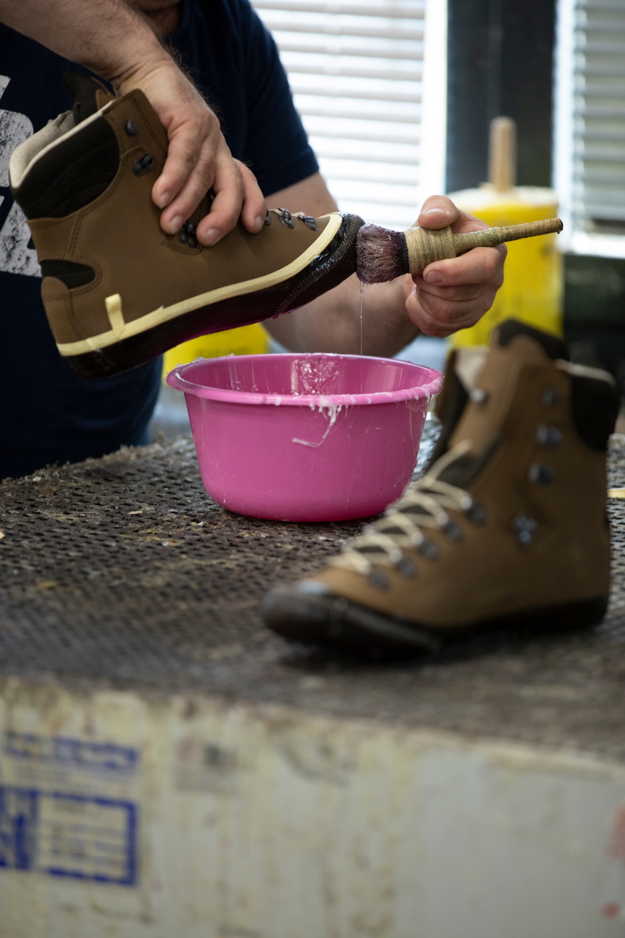 Ein Mitarbeiter streicht mit einem Pinsel Kleber auf die Sohle eines Bergschuhs, daneben steht eine pinkfarbene Schüssel