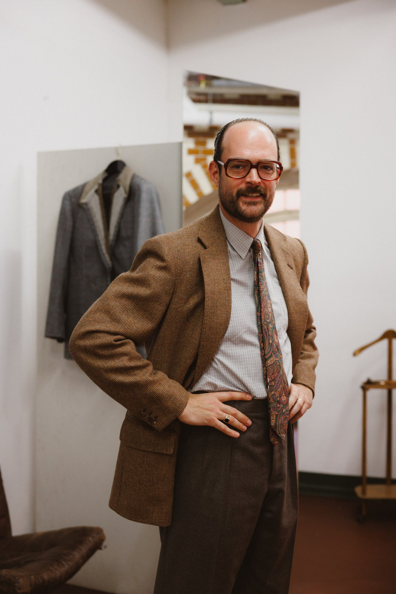Mann im braunen Tweedsakko mit Paisley-Krawatte, eine Hand in der Hüfte, in einem weißen Raum, hinter ihm ein graues Sakko