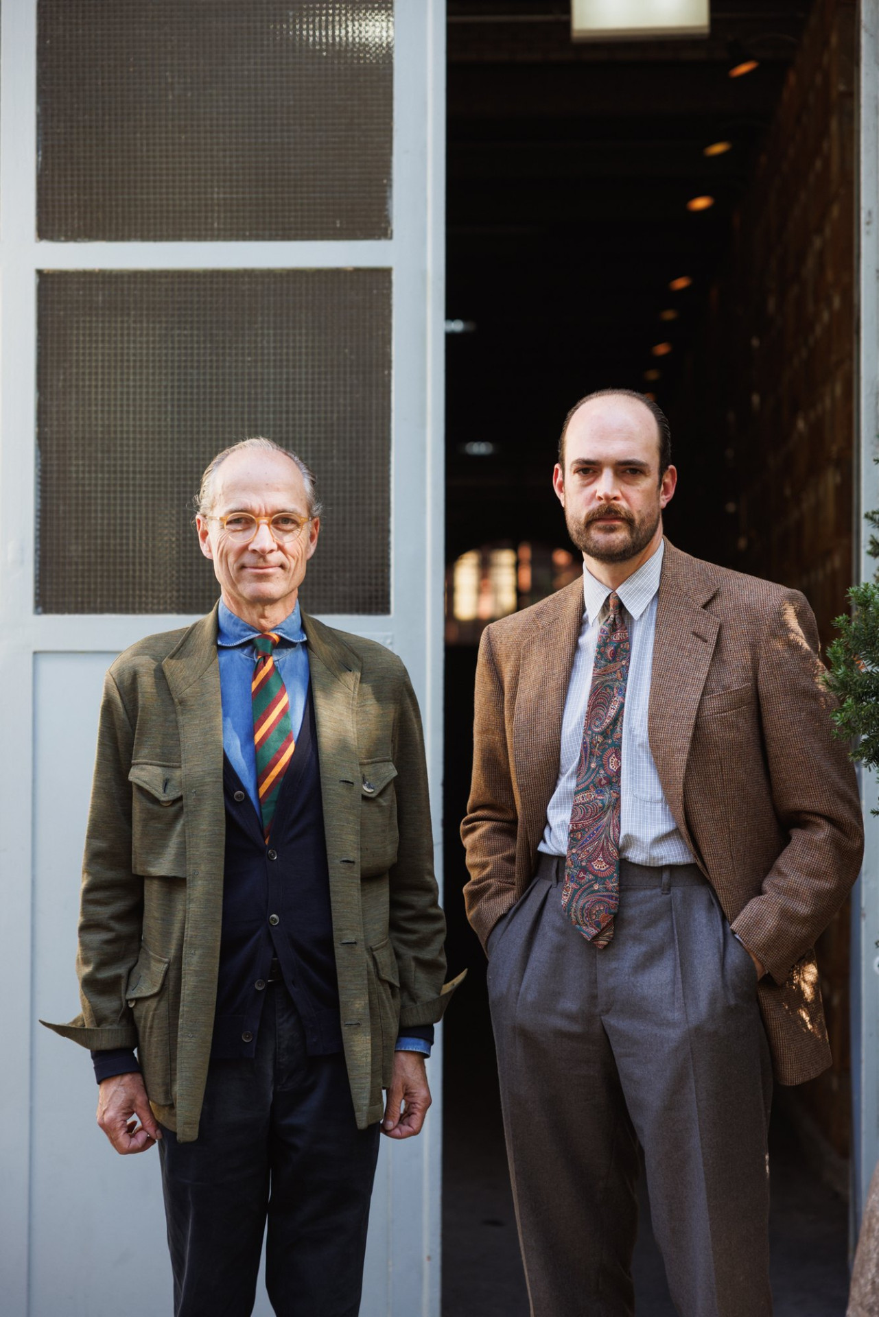 Zwei Männer stehen vor einer Hoftür, links in grüner Jacke und dunkelblauer Weste, rechts im braunen Tweedsakko mit grauer Hose